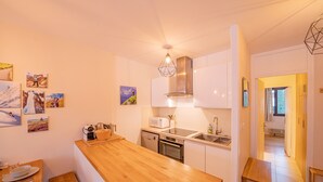 Una cocina compacta con armarios blancos, encimera de madera, microondas, cocina, y un lavabo. Hay cuadros en la pared y un taburete de madera pequeño.
