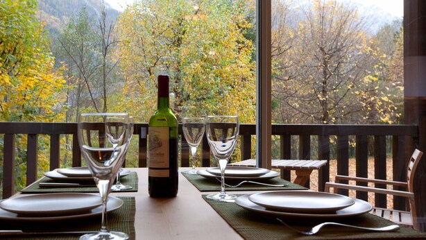 Una mesa de comedor dispuesta con platos, vasos y una botella de vino, con vistas a un paisaje montañoso con follaje otoñal.