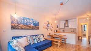 Un salón acogedor con un sofá azul, suelo de madera y una zona de cocina con armarios blancos y una mesa de madera.