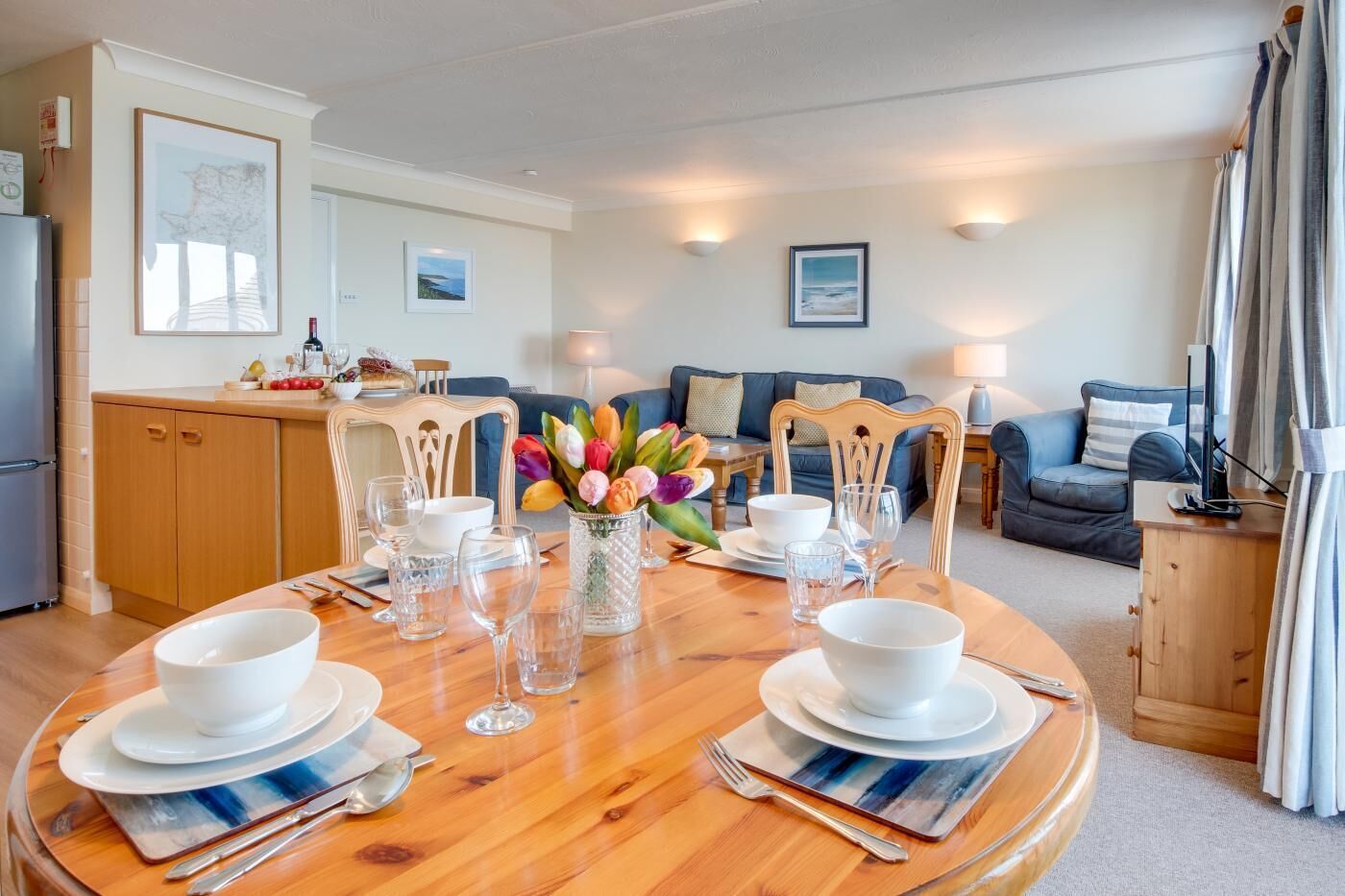Un comedor con una mesa redonda dispuesta para cuatro personas, una cocina al fondo y una sala de estar con un sofá y lámparas.