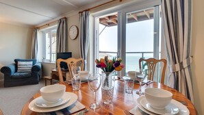 A dining area with a wooden table set for four, a sofa, a TV, and a view of the sea. - 2 Bed in Clifton Court (oc-fl24) (Croyde)