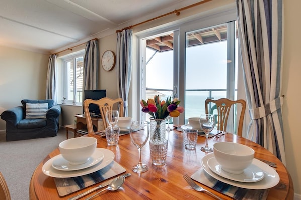 A dining area with a wooden table set for four, a sofa, a TV, and a view of the sea.