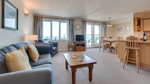 A living room with a sofa, a coffee table, and a TV. There is a dining area with a table and chairs. The room has large windows with curtains and a view of the sea. - 2 Bed in Clifton Court (oc-fl24) (Croyde)