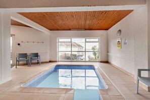 Una piscina interior con techo de madera, dos sillas y un ventanal con vista a una casa.