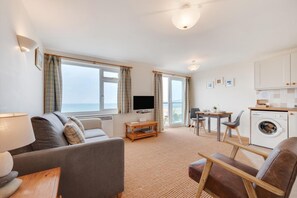 A modern living room with a sofa, a TV, a dining area, and a kitchen with a washing machine. - 1 Bed in Clifton Court (oc-fl30) (Croyde)