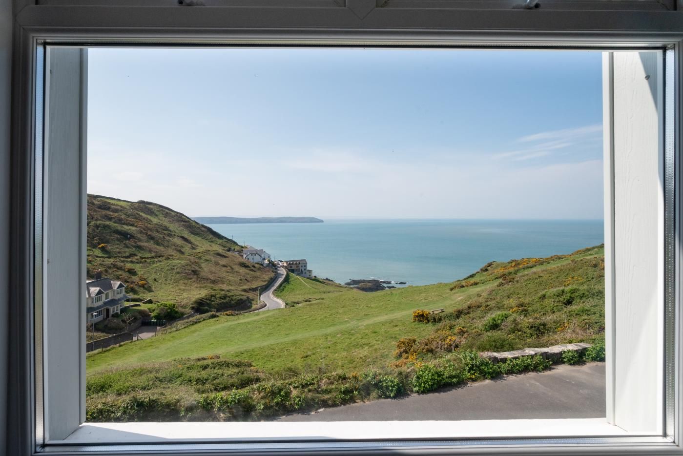 A coastal view from a window, featuring a road leading to houses on a hillside, overlooking the sea.