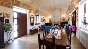 Un comedor con una mesa de madera larga, sillas y un banco. Hay dos ventanas con cortinas, una chimenea y luces fijadas a la pared.