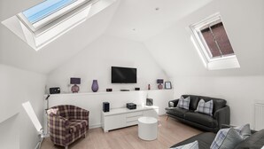 Ein modernes Wohnzimmer im Loft-Stil mit Flachbildfernseher, weißem Konsolentisch, schwarzem Ledersofa, Ohrensessel mit Karomuster und einem Oberlicht.