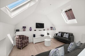 A modern loft-style living room with a flat-screen TV, a white console table, a black leather sofa, a plaid armchair, and a skylight. - Historic 2‑Bedroom Central St Andrews Cottage Near West Port, Stylish Interiors & Walk to Old Course (St. Andrews)