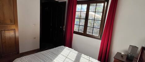 Un dormitorio con una cama grande, vigas de madera en el techo, un ventanal con cortinas rojas y una mesita de noche con una lámpara.