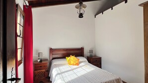 A bedroom with a wooden ceiling, a bed with a white bedspread, a nightstand with a lamp, and a red curtain on the window.