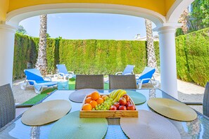 An outdoor dining area with a table set for two, surrounded by blue lounge chairs, and a fruit basket on the table.