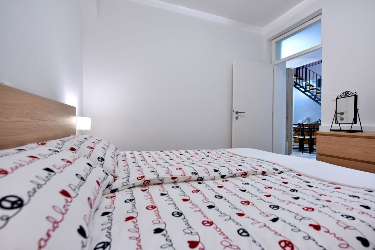 A bedroom with a neatly made bed, a wooden headboard, a bedside table, and a view of a staircase leading to a balcony.