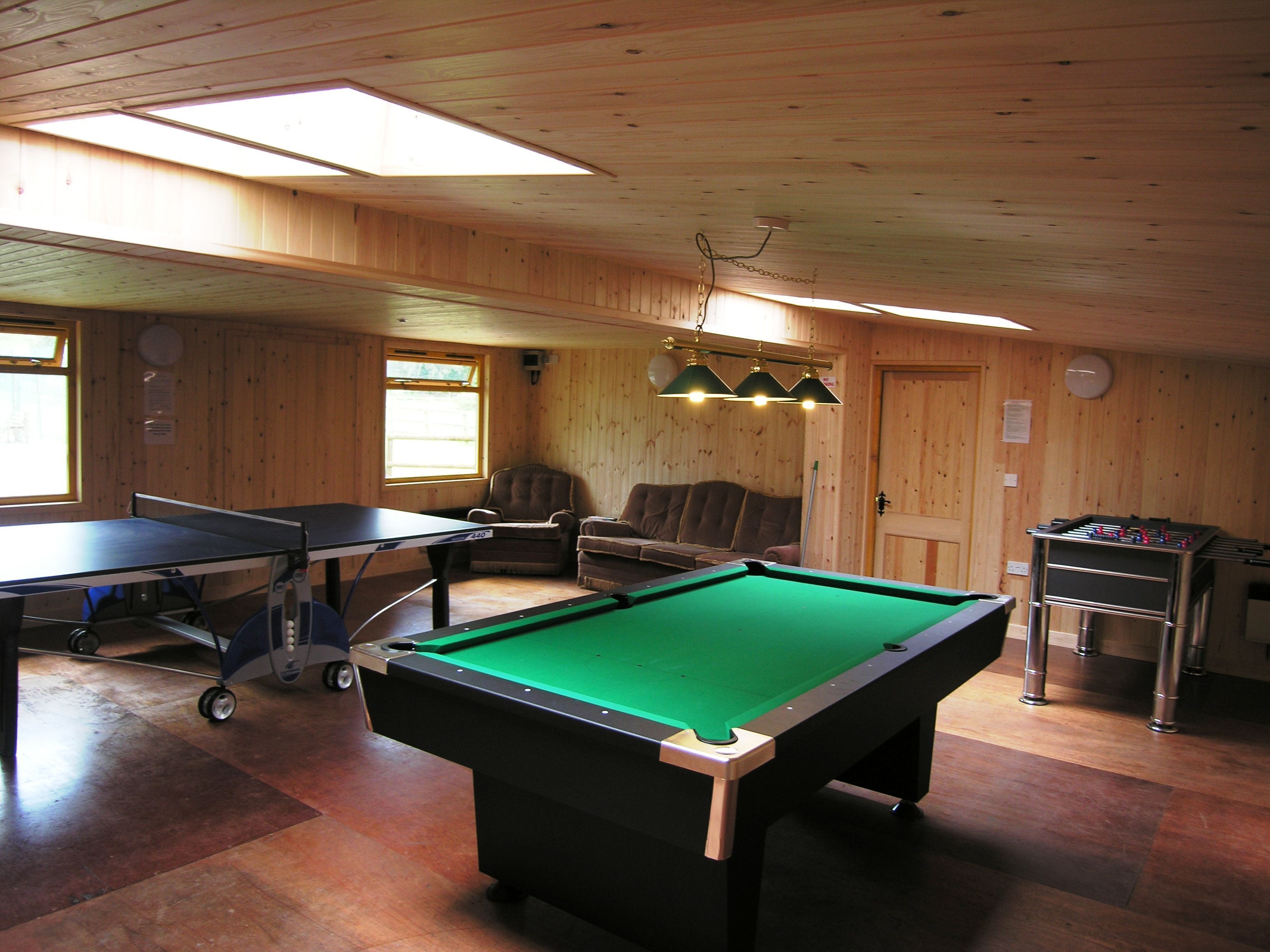 Une table de billard recouverte d’un drap vert, une table de baby-foot et une table de billard dans une pièce aux murs et au plafond en bois.