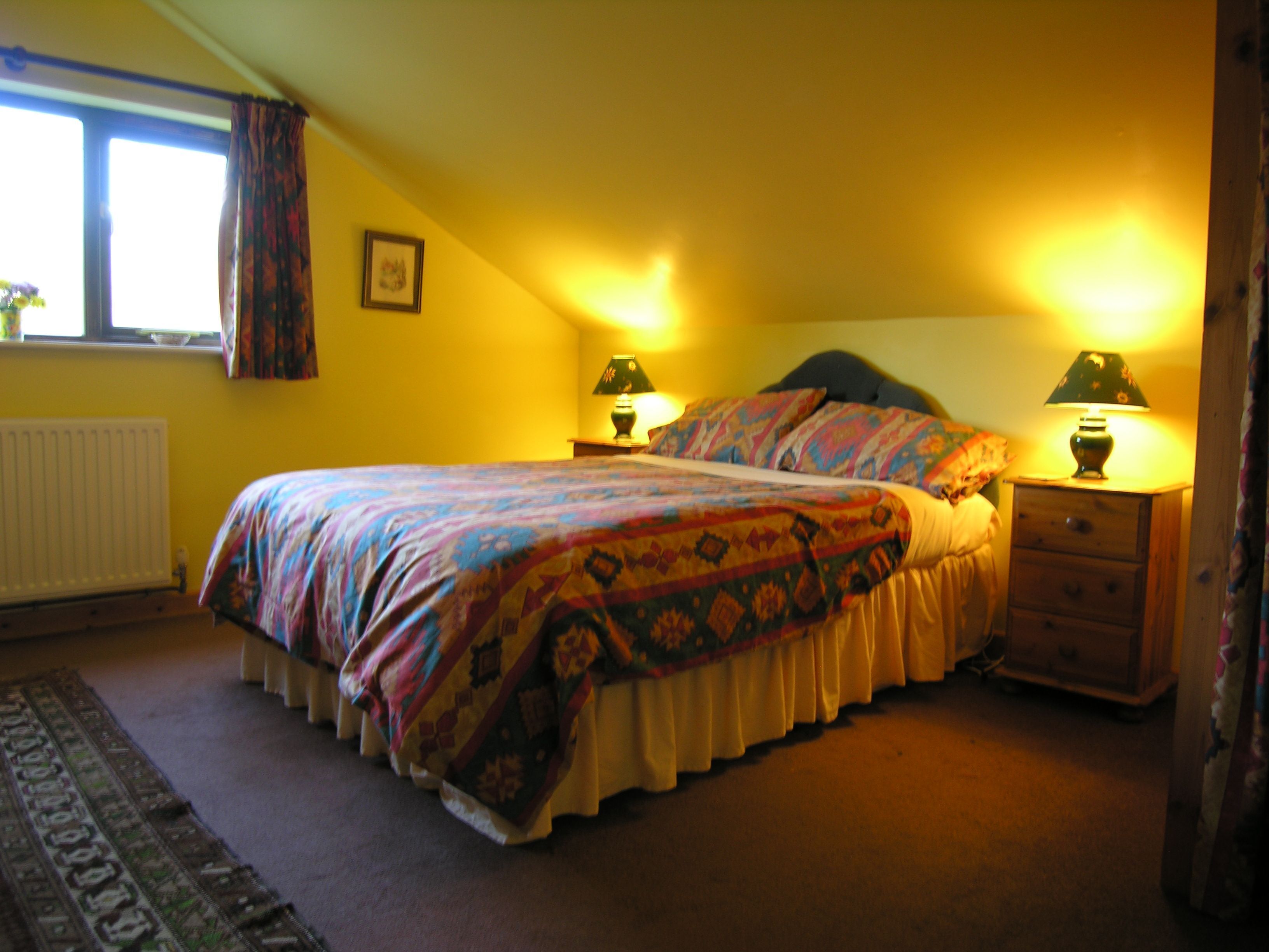 Een slaapkamer met een bed, twee nachtkastjes met lampen, een radiator, een raam met gordijnen en een vloerkleed.