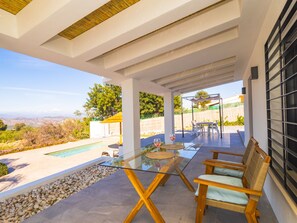 Une terrasse extérieure couverte, dotée d’une table en verre, de chaises en bois, d’une piscine et offrant une vue sur le paysage.