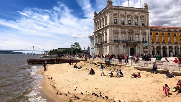 Una playa con gente y palomas, un puente al fondo y edificios históricos.