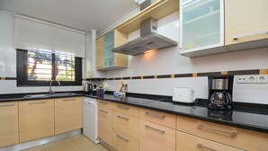 A modern kitchen with light wood cabinets, a black countertop, and a window with blinds. - Jardines del Pla - Javea, Costa Blanca (Javea)