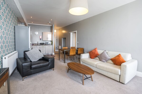 Un salon moderne avec un canapé en cuir noir, un canapé blanc agrémenté de coussins orange, une table basse en bois et un coin repas comprenant une table et des chaises.