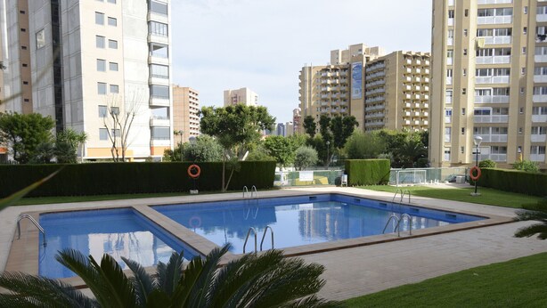 A swimming pool surrounded by well-maintained grass and trees, with tall residential buildings in the background. - PARAISO ESPERANTO (1 BEDROOM) (Benidorm)