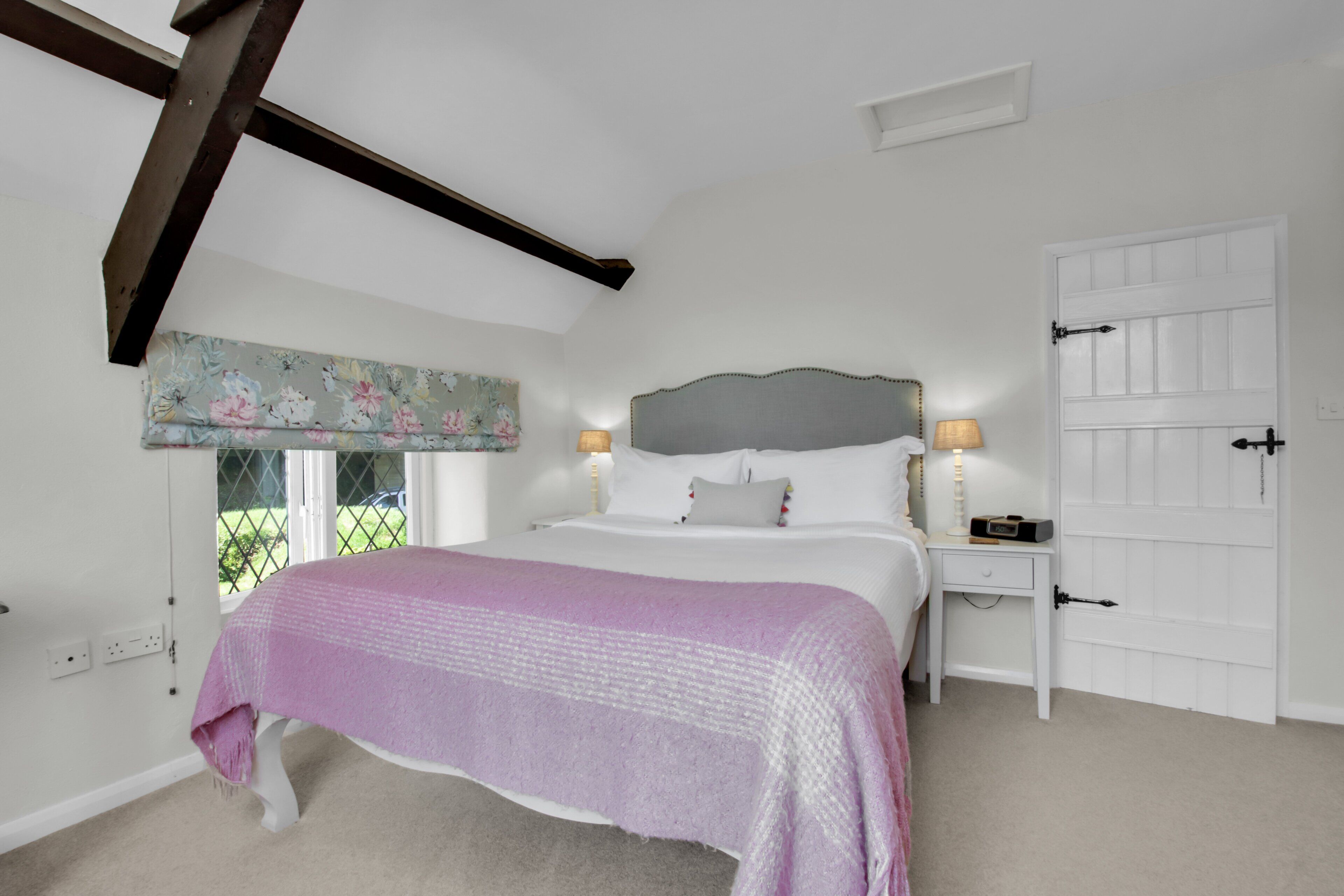 A bedroom with a bed, bedside lamps, a window with a floral curtain, and a white door.