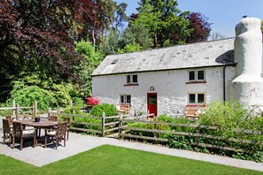 A white stone house with a red door, a wooden fence, and a garden with a table and chairs. - Cascade Cottage, Exford (Exford)