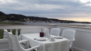 A table set for two with white chairs and a tablecloth, overlooking a body of water and a hillside with buildings. - Classic attic apartment (Supetarska Draga)