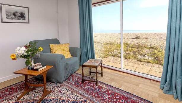 Een woonkamer met een grijze bank, een bijzettafeltje met een bloevas, een kamerkleed met patroon en uitzicht op een rotsachtig landschap door grote ramen.