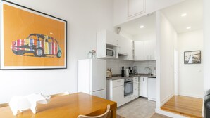 Une cuisine moderne avec des meubles blancs, une table à manger en bois et un tableau coloré représentant une voiture accroché au mur.