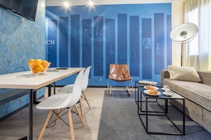 Ein modernes Wohnzimmer mit Sofa, Couchtisch, Esstisch mit Stühlen, einem an der Wand befestigten Fernseher und einem großen Fenster mit Vorhängen.