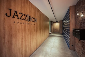 Passadís amb paret de fusta que mostra el rètol 'JAZZBCN APARTMENTS', un paviment de formigó i una paret de maó amb una prestatgeria metàl·lica a la dreta.