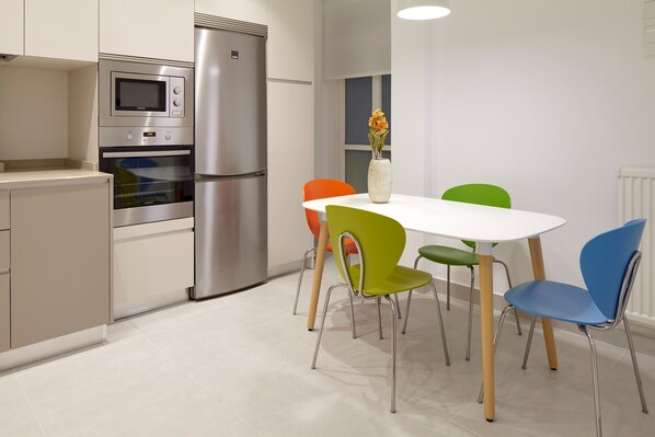 A modern kitchen with stainless steel appliances, a white dining table, and colorful chairs.