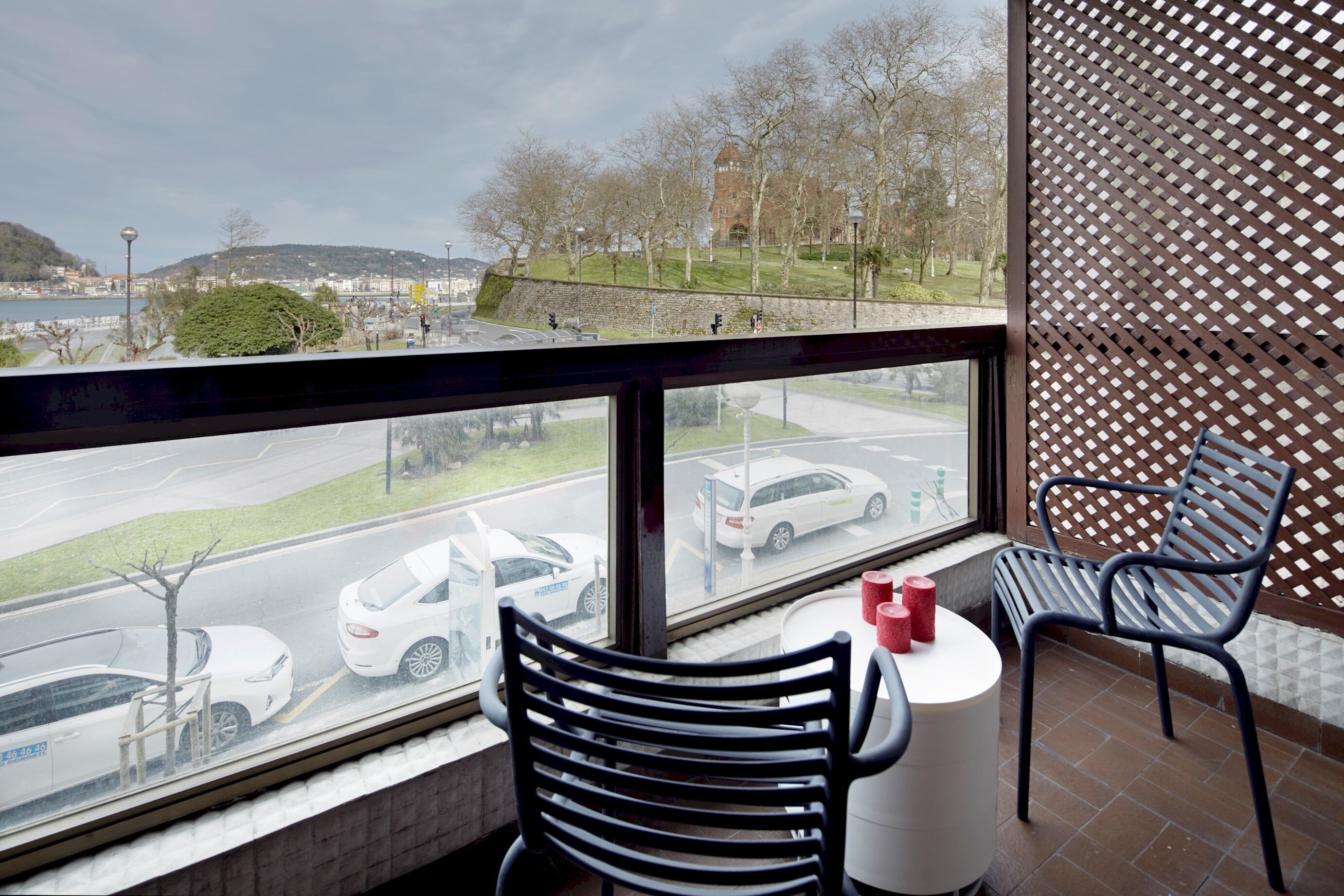 A balcony with a view of a road and parked cars, two chairs, and a small table with candles.