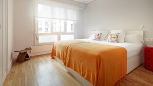 Un dormitorio con un ventanal grande, una cama con ropa de cama blanca y un plaid naranja, una mesita de noche roja y suelo de madera.