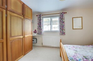 A bedroom with wooden wardrobe, a bed with floral bedding, a window with floral curtains, and a radiator.
