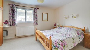 A bedroom with a wooden bed, a nightstand, a bedside table, a window with curtains, a radiator, and a ceiling light fixture. - 1 Bed in Ilfracombe (oc-n2052) (Berrynarbor)