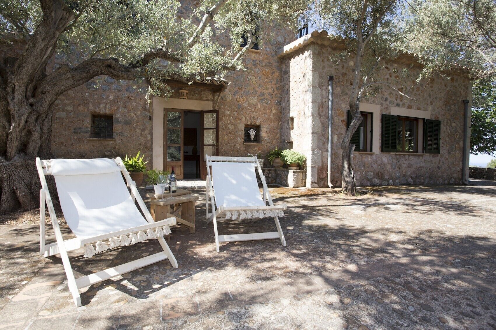 A stone building with a rustic exterior, two white lounge chairs, and a small table with a bottle and a plant.