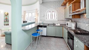 Une cuisine moderne avec des meubles de couleur verte, un four encastré, un four à micro-ondes et une machine à café. On y trouve deux chaises bleues et un petit îlot central avec un évier.