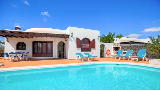 Un espace aménagé au bord d’une piscine à l’eau d’un bleu limpide, avec un bâtiment blanc doté de fenêtres en arc de cercle et des chaises longues bleues.