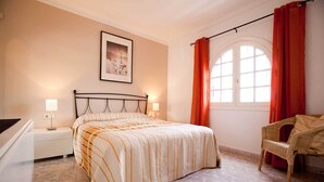Une chambre à coucher comprenant un lit, des tables de chevet, une chaise, une fenêtre avec des rideaux et un tableau encadré accroché au mur.