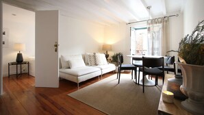 Ein modernes Wohnzimmer mit einem weißen Sofa, einem Esstisch mit Stühlen, einem kleinen Beistelltisch mit einer Lampe und einem großen Fenster mit Vorhängen.