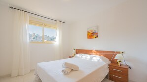 A neatly made bed with white linens and a rolled towel, a wooden nightstand with a lamp, a window with white curtains, and a painting on the wall. - Apartment Zeus - PlusHolidays (Calpe)