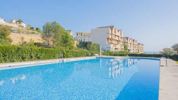 A swimming pool with a clear blue water surface, surrounded by greenery and residential buildings. - Apartment Zeus - PlusHolidays (Calpe)