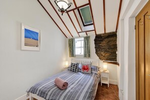 Ein Schlafzimmer mit Bett, Nachttisch, Lampe, einem Bild an der Wand, einem Fenster mit Vorhängen und einer Steinwand.