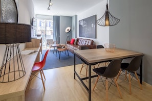 A modern living room with a dining area, featuring a wooden dining table, red chairs, a brown leather sofa, and a large window with a view of buildings.
