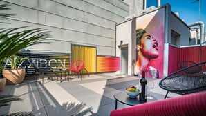 An outdoor patio area with a modern design, featuring a seating area with chairs and a table, a potted plant, and a wall with a colorful mural.