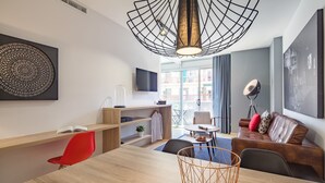 A modern hotel room with a wooden desk, red chair, and a dining table. There is a sofa with cushions, a floor lamp, and a large window with a view of buildings.