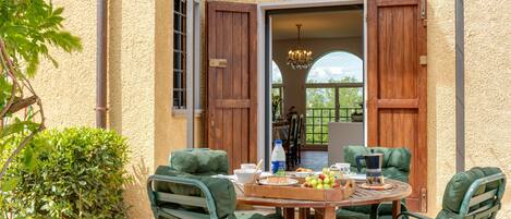 An outdoor patio with a wooden table, green cushioned chairs, and a view of the interior.
