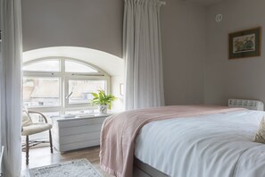 A bedroom with a large bed, a desk with a plant, a chair, and a window with curtains.