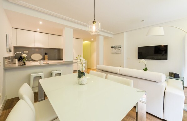 Una cocina moderna con armarios blancos, un comedor con una mesa y sillas blancas, y una sala de estar con un sofá blanco y un televisor de pantalla plana.